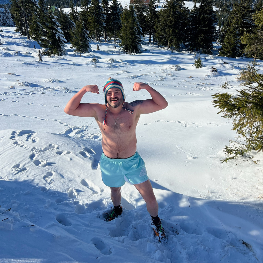 Ricardo Castro Ramírez posando sonriente y mostrando fuerza en un entrenamiento de exposición al frío, de pie en la nieve con shorts y gorro de lana, rodeado de un paisaje nevado y árboles.