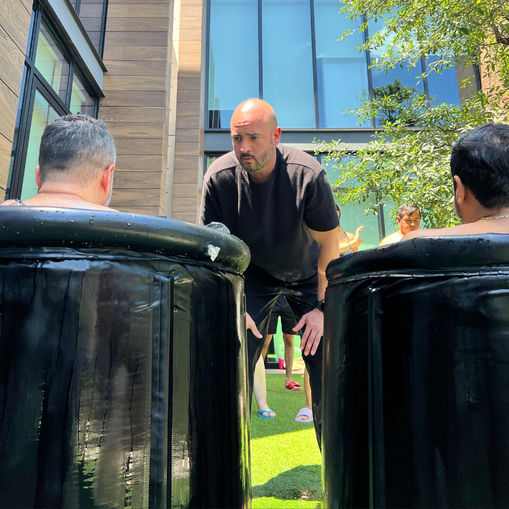 Ricardo Castro Ramírez guiando a dos participantes durante una sesión de inmersión en hielo, observándolos con atención mientras están en contenedores negros con agua fría, en un entorno al aire libre.