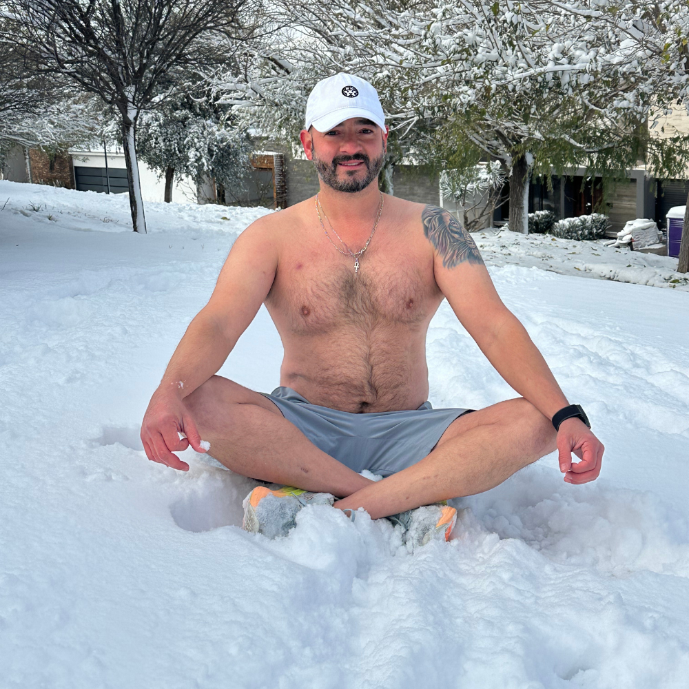 Ricardo Castro Ramírez sentado en posición de meditación sobre la nieve, sin camiseta y con gorra blanca, practicando respiración y conexión con el frío.