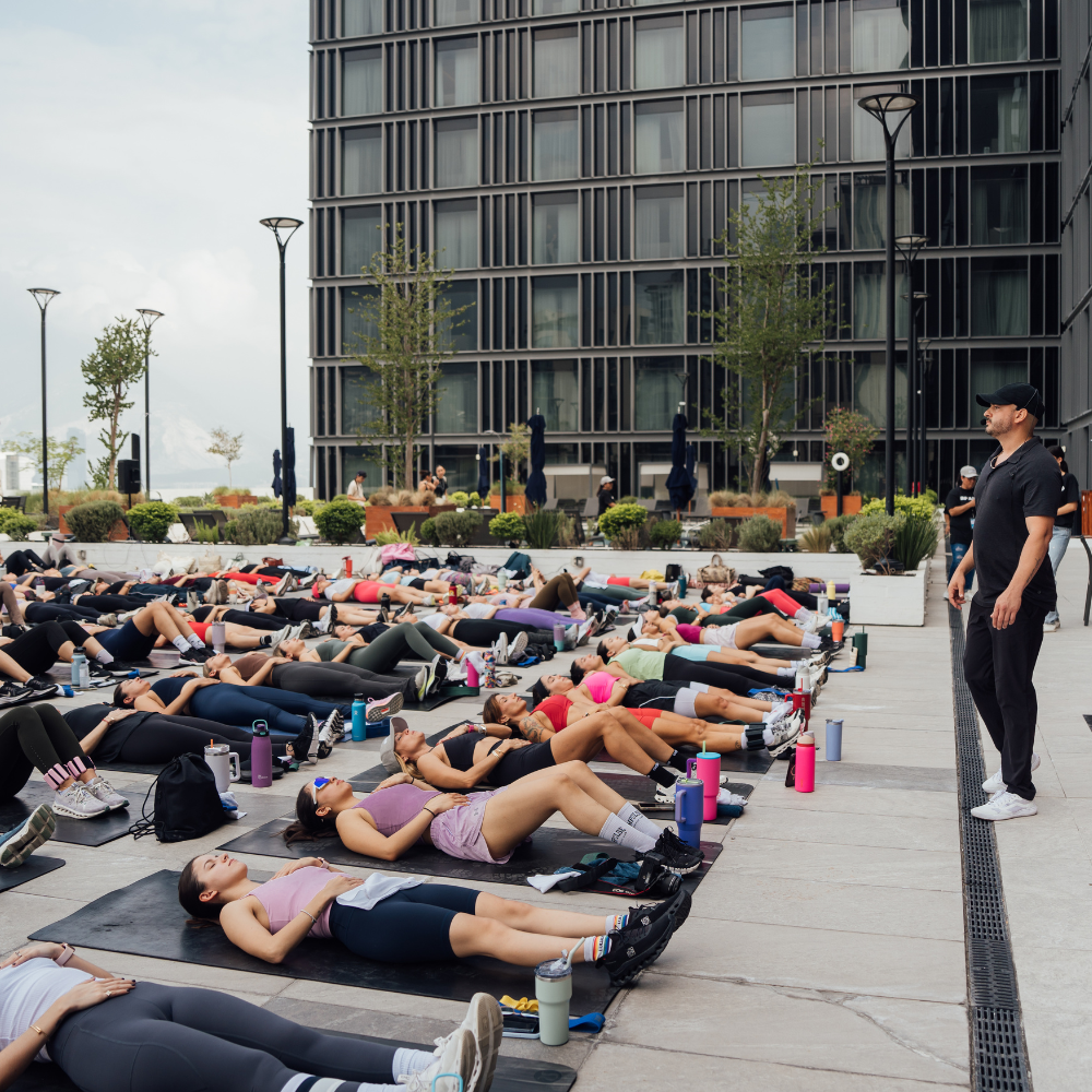 Richi Castro guiando una sesión de breathwork al aire libre, con decenas de personas recostadas en tapetes frente a un edificio moderno.